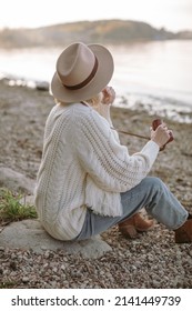 A Traveler Dressed In Cowboy Style Sitting On The Shore And Taking A Pictures Of A Sunset On Her Film Camera.