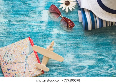 Traveler Essentials On Blue Wooden Background- A Wooden Toy Airplane, A Beach Bag,a White Hat And A Folded Map. Space For A Text Or Product Display, Top View.