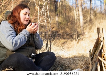 Traveler fat woman wearing casual clothes sitting with cross legs in the forest near the bonfire holding in her hands a cup of hot tea.