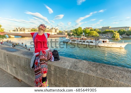 Traveler freedom concept. Caucasian lifestyle woman with open arms enjoying the Seine. Bateau-mouche and Pont Neuf on a blurred background. Tourist traveler and popular landmarks of Paris, France.