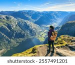 A traveler gazes at the stunning fjord landscape, bathed in early morning light, surrounded by towering mountains and lush valleys. woman with backpack hiking Kjeragbolten Lysefjord in Norway