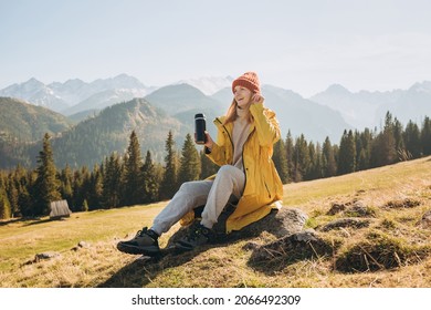 Traveler girl drinking tea from thermos cup over nature background. Mountain peaks. Freedom, happiness, travel and vacations concept, outdoor activities, woman wearing red hat
