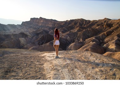 Traveler girl with fit body is standing in white short shorts in front of dry hot lifeless desert landscape, Death Valley National Park, California, back of red long hair woman in nature, Holiday trip