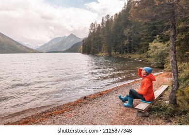 A Traveler Girl Is Sitting On A Bench And Enjoying A Great Panoramic View Of A Mountain Lake In Altai. Hiking In The National Park And Solitude In Nature