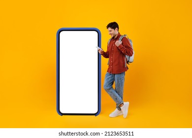 Traveler Guy Using Mobile Phone Standing Near Big Cellphone With Blank Screen Standing With Backpack On Yellow Studio Background. Travel Application For Your Smartphone. Mockup