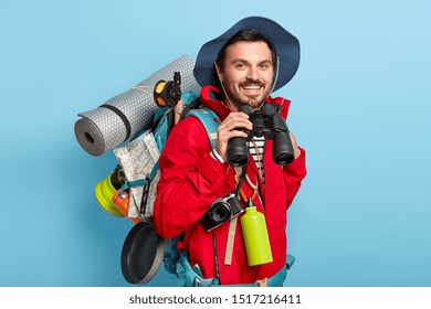Traveler man has walk with backpack, carries necessary things for trip, looks through binoculars, has glad look, wears casual clothes, poses against blue background. People, journey, lifestyle concept