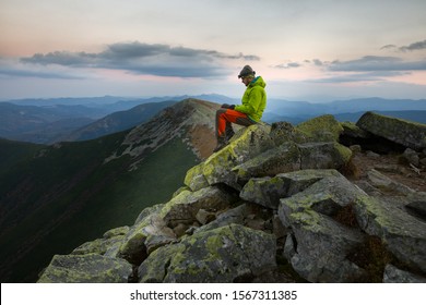 Traveler Man In A Hat With A Headlamp Sits On Top Of A Stony Mountain In The Evening And Looks Down.