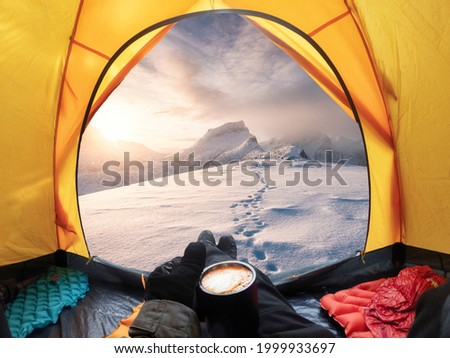 Traveler man holding coffee cup and enjoying the view of sunrise on snowy mountain inside a yellow tent