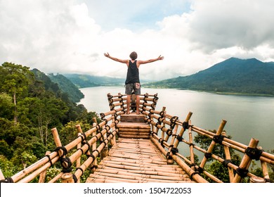 Traveler Man Standing On Two Steps Of A Wooden/bamboo Balcony In Bali. Rear View Of Guy Enjoy Looking Picturesque View Of Mountains And Water In Indonesia. Concept Of Real Adventure Holiday Vacation. 