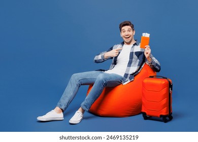 Traveler man wear casual clothes sit in bag chair near suitcase hold point finger on passport ticket isolated on plain blue background. Tourist travel abroad in free time getaway. Air flight concept - Powered by Shutterstock