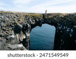 Traveler on a Stone bridge. Gatklettur Arch Rock cliff with natural arch near Arnarstapi, Snaefellsnes, West Iceland. Famous basalt Stone Arch. Famous tourist 