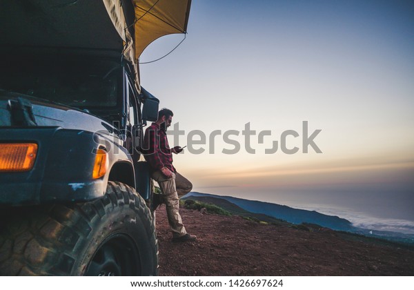 Traveler people with car and camping concept -\
lonely man use cellular phone to connect to internet outside his\
vehicle - mountain and nature outdoor around - enjoying freedom and\
alternative vacation