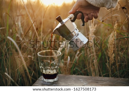 Traveler pouring fresh hot coffee from geyser coffee maker into glass cup in sunny warm light in rural countryside herbs. Atmospheric tranquil moment. Alternative coffee brewing in travel