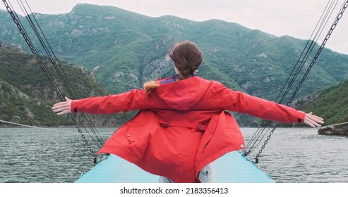 Traveler In Red Jacket Raises Arms To Sides On Moving Boat, Background Of Mountains Landscape. Woman Travel By Ferry On Albania Lake. Adventure, Vacation Concept. Explore Beauty World, Recreation
