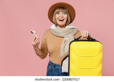 Traveler Tourist Amazed Mature Elderly Senior Lady Woman 55 Years Old Wears Brown Shirt Hat Scarf Hold Suitcase Bag Use Mobile Cell Phone Isolated On Plain Pastel Light Pink Background Studio Portrait