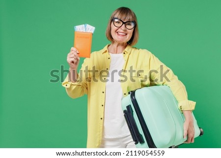 Traveler tourist elderly fun woman in yellow shirt glasses hold suitcase valise passport ticket isolated on plain green background Passenger travel abroad on weekend getaway Air flight journey concept