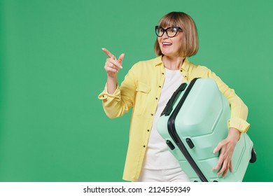 Traveler tourist elderly woman in yellow shirt glasses hold suitcase valise point finger aside isolated on plain green background Passenger travel abroad on weekends getaway Air flight journey concept