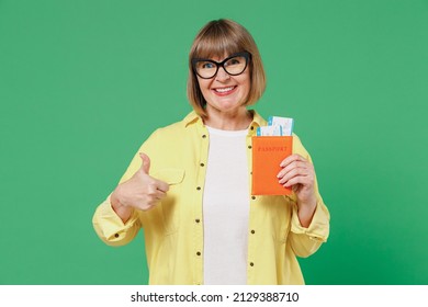 Traveler tourist elderly woman in yellow shirt glasses hold passport tickets show thumb up isolated on plain green background studio. Passenger travel abroad on weekends. Air flight journey concept