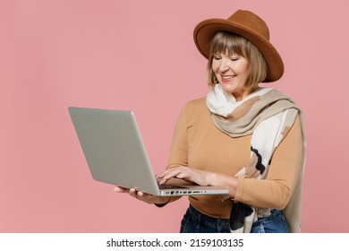 Traveler Tourist Excited Mature Elderly Senior Lady Woman 55 Years Old Wears Brown Shirt Hat Scarf Hold Use Work On Laptop Pc Computer Isolated On Plain Pastel Light Pink Background Studio Portrait