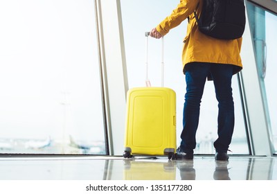 Traveler Tourist Hold Yellow Suitcase Backpack Is Standing At Airport On Background Large Window, Man In Bright Jacket Waiting In​ Airport Lobby Terminal, Vacation Trip Concept, Empty Space Mockup