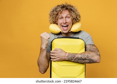 Traveler Tourist Man He Wear Grey T-shirt With Neck Pillow Hold Suitcase Do Winner Gesture Isolated On Plain Yellow Background. Passenger Travel Abroad On Weekends Getaway. Air Flight Journey Concept.