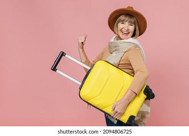 Traveler tourist mature elderly senior woman 55 years old wears brown shirt hat scarf hold under hand suitcase bag clench fists say yes isolated on plain pastel light pink background studio portrait