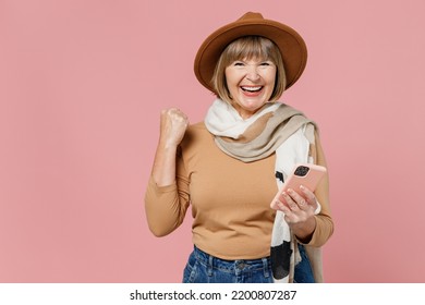 Traveler Tourist Mature Elderly Senior Lady Woman 55 Years Old Wears Brown Shirt Hat Scarf Hold Use Mobile Cell Phone Clench Fist Say Yes Isolated On Plain Pastel Light Pink Background Studio Portrait