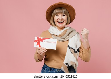 Traveler tourist winner elderly senior woman 55 years old wears brown shirt hat scarf hold gift certificate coupon voucher card for store isolated on plain pastel light pink background studio portrait