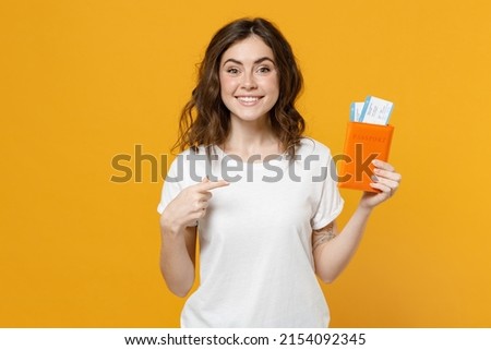 Traveler tourist woman 20s in white t-shirt point index finger on passport with tickets isolated on yellow orange background. Passenger traveling abroad on weekends getaway. Air flight journey concept