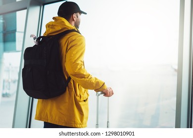 Traveler Tourist With Yellow Backpack Is Standing At Airport On Background Large Window, Man In Bright Jacket Waiting In  Hall Of ​ Airport Lobby Terminal, Vacation Trip Concept, Empty Space Mockup 