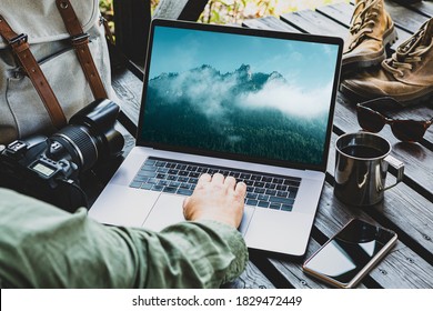 Reisende oder Reisefotograf, die auf Laptop-Computer im Freien arbeiten, unter seiner Ausrüstung im Hintergrund (Rucksack, Kamera, Smartphone, Stiefel). Berglandschaftsfoto auf dem Bildschirm.