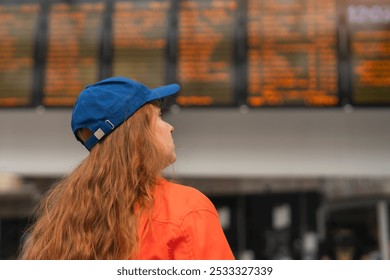 A traveler in a vibrant orange jacket gazes at a bustling train station schedule board on a busy afternoon - Powered by Shutterstock