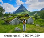 A traveler wanders towards charming turf-roofed houses beneath a towering mountain in Norway