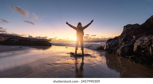 Traveler Woman with arms raised in triumph on a beach at sunset, silhouetted against vibrant sky and crashing waves. Travel and Freedom Concept image - Powered by Shutterstock