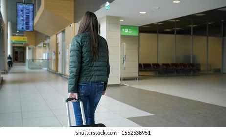 Traveler Woman Brunette Is Walking In Modern Airport Terminal Carrying Suitcase, Back View. Passenger Is Going To Her Flight In City Airport.