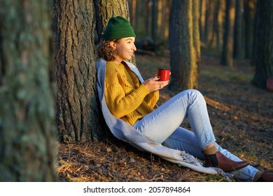 Traveler woman enjoy hot tea in forest. Young female hiker relaxing sit at tree drink hot beverage from metal mug happy smiling look at beautiful sunset after wondering in autumn woods all day long