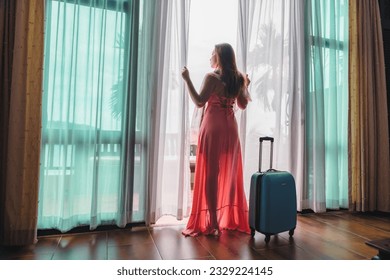 Traveler woman in pink dress with suitcase standing in room her hand opening curtains looking out of full-length window of modern luxury resort or hotel, Lifestyle tourist girl on holiday vacation