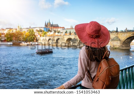 A traveler woman with a sunhat enjoys the view over the Vlatava River to the Charles Bridge and castle of Prague, Czech Republic, during a sunny autumn day