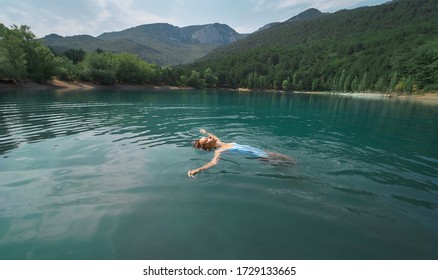 Traveler Woman Swimming On Back In Beautiful Mountain Lake With Clear Blue Water Against Mountains. Beautiful Scenery, People In Nature And Travel Concept