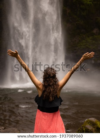 Traveler woman wearing pink dress at waterfall. Excited woman raising arms in front of waterfall. Travel lifestyle. View from back. Copy space. Nung Nung waterfall in Bali, Indonesia