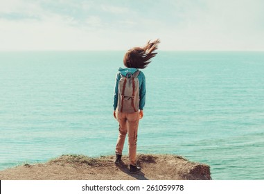 Traveler young woman with backpack standing on coastline near the sea in windy weather, her hair fluttering in the wind