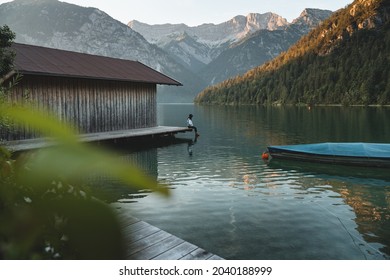 Traveler young woman relaxing on pier in Plansee Lake. Hipster female with denim jacket and a hat sitting on wooden pier and looking at view. Vacation concept. Austria,Tirol.