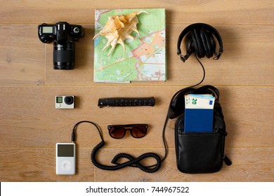 A Traveler's Kit. 
Photo And Action Cameras, Music Player, Sunglasses, Headphones, Document Bag With Passport And Money Inside, Pocket Knife, Paper Map And A Sea Shell.
Wooden Background.
Top View.