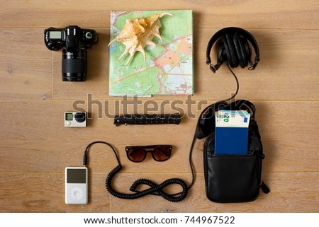 A traveler's kit. 
Photo and action cameras, music player, sunglasses, headphones, document bag with passport and money inside, pocket knife, paper map and a sea shell.
Wooden background.
Top view.