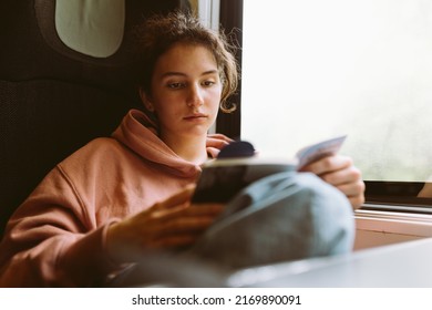 Traveling by train of lonely young woman. teenage girl in sweatshirt, sits by window in train car, reads book, dreams alone, spends time on road - Powered by Shutterstock