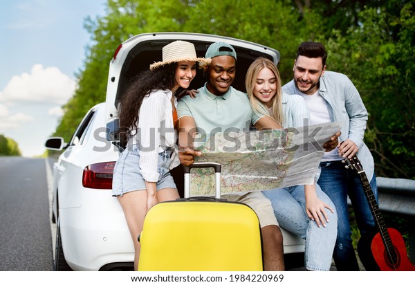 Traveling\
with friends concept. Happy multiracial young people sitting in\
open car trunk, checking map, finding their travel destination,\
having fun road trip adventure in\
summertime