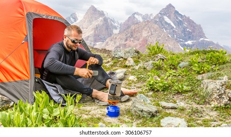 Traveling Man Eating Meal.
Hiker Sitting In His Orange Camping Tent And Having Lunch Stove And Cooking Gear Mountain Landscape On Background