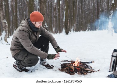 Traveling man a sits near camp fire and warm his hands. Winter time. Concept adventure active vacations outdoor. Extreme camping. vintage backpack and thermos with tea, snowy forest