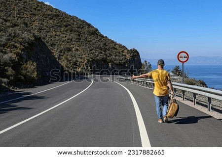 Traveling musician with a guitar walking alone down the winding road with a beautiful sea view, trying to stop a car by showing a hitchhiking gesture.