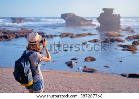Traveling and photography. Young woman with camera and backpack taking picture on the sea beach.